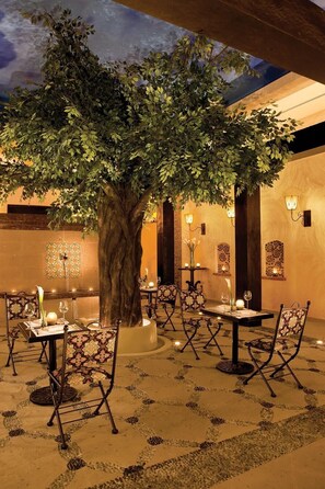 An outdoor dining area with tables and chairs, a tree, and wall-mounted lights. - All inclusive romantic swim out room with jacuzzi for 2 at Secrets Maroma Beach (Playa del Carmen)