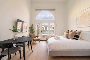 A modern bedroom with a large bed, a dining table, and a chair. There is a window with a view of buildings outside.