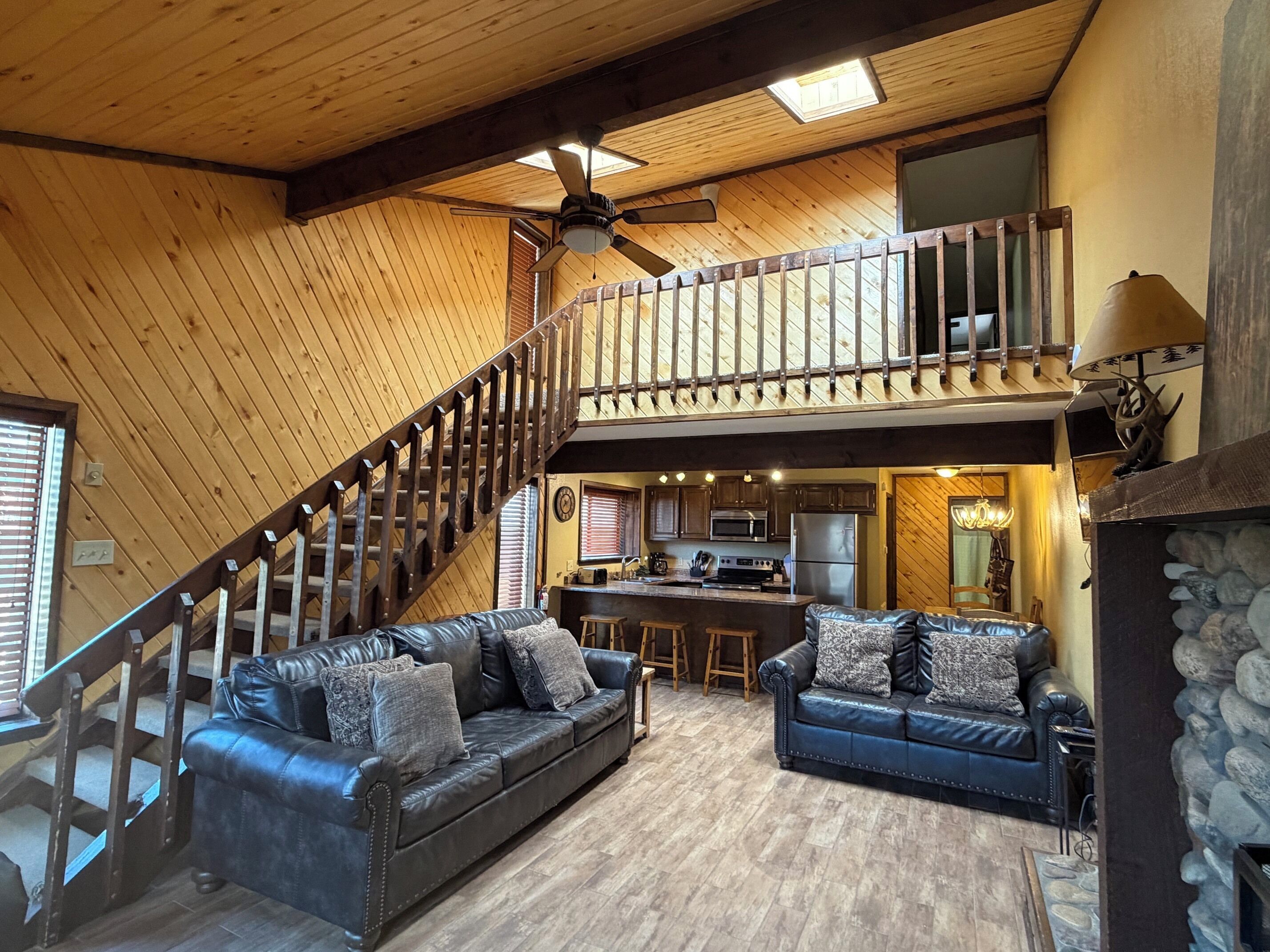 A spacious living area with a high ceiling, a wooden staircase, a kitchen area, and a seating arrangement with two sofas.