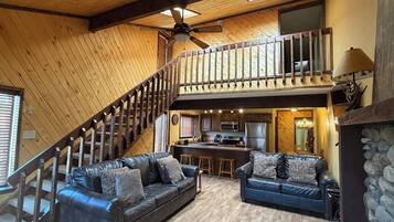 A spacious living area with a high ceiling, a wooden staircase, a kitchen area, and a seating arrangement with two sofas.