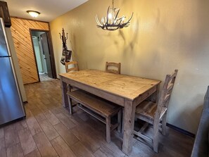 A dining area with a wooden table and chairs, a chandelier, and a refrigerator.