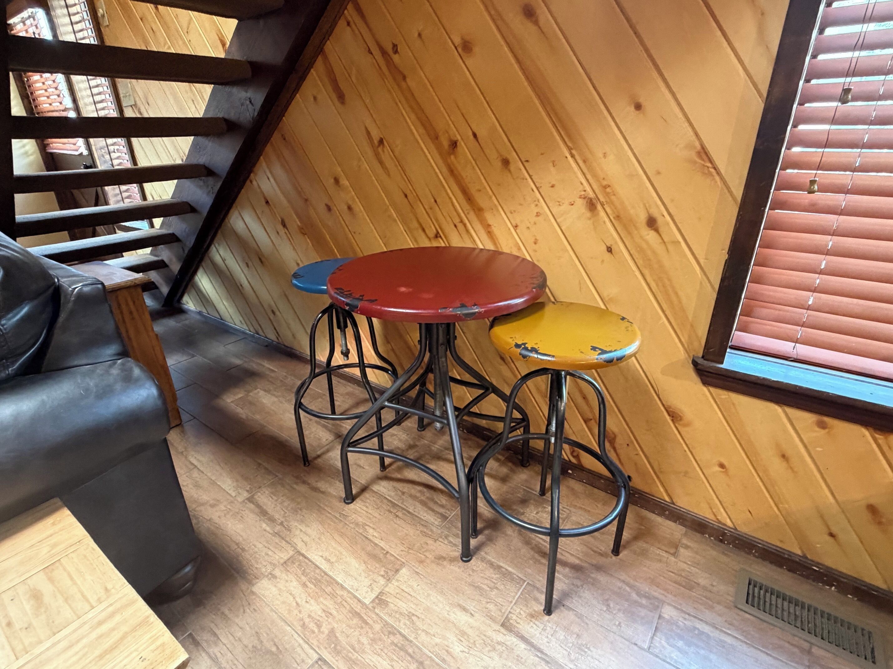 A corner of a room with a wooden sloped ceiling, a black leather sofa, and a small round table with stools.