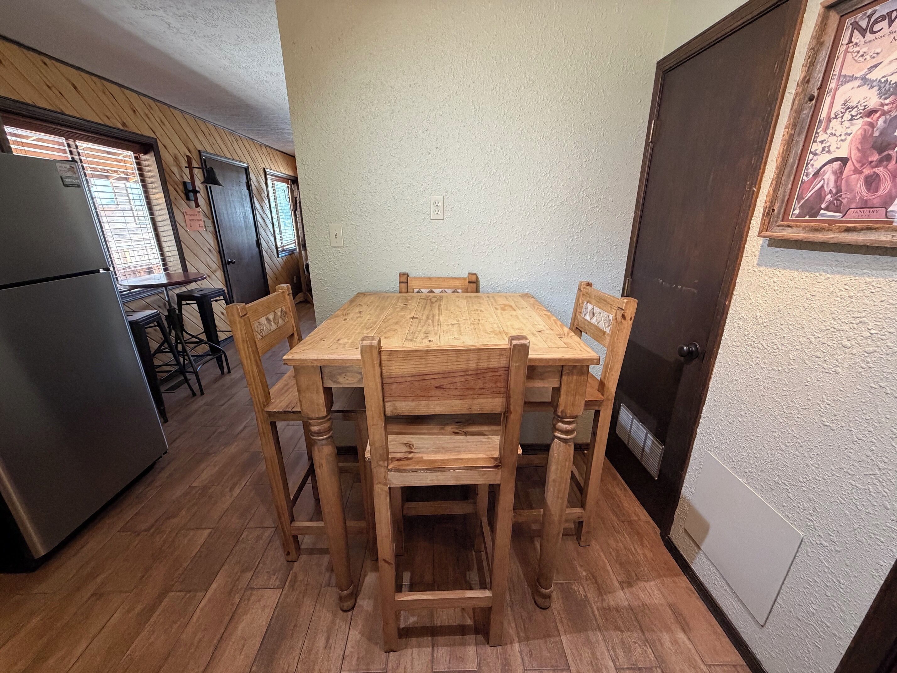 Une table de salle à manger en bois, quatre chaises, un réfrigérateur et un plan de travail de cuisine avec un évier.