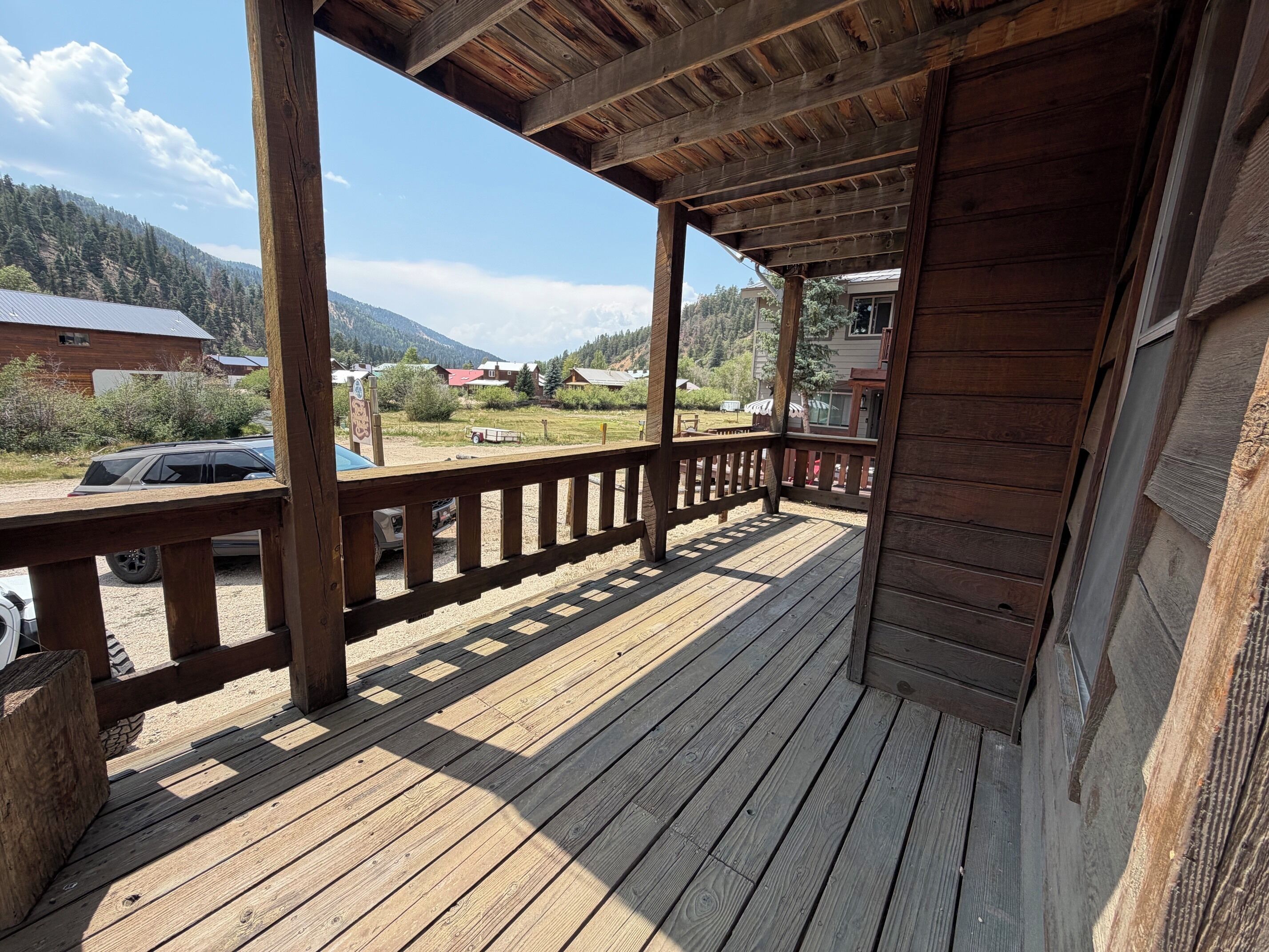 Terraza de madera con barandilla, con vista a un estacionamiento y un paisaje montañoso.