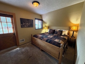 Un dormitorio con cama de madera, dos ventanas con persianas, una lámpara de techo y una mesita de noche con lámpara.