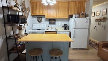 Una cocina con armarios de madera, una isla central con taburetes, microondas, estufa, refrigerador y una araña.