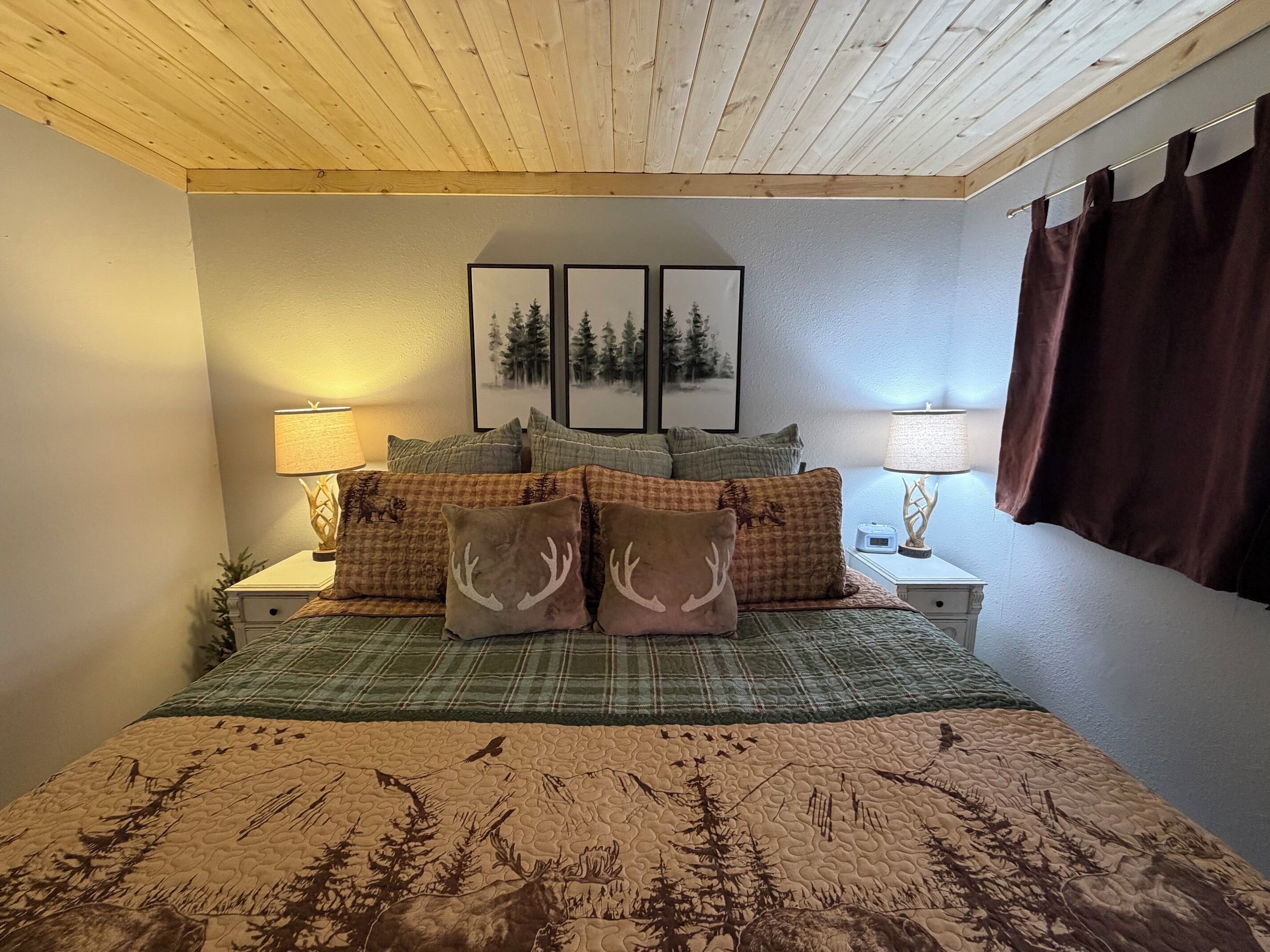 A bedroom with a wooden ceiling, a bed with a forest-themed quilt, two bedside lamps, and a window with curtains.