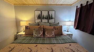 A bedroom with a wooden ceiling, a bed with a forest-themed quilt, two bedside lamps, and a window with curtains.