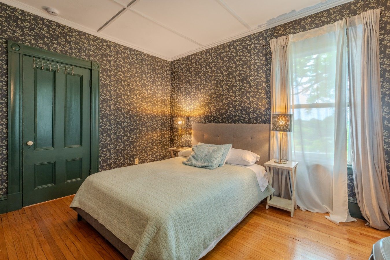 A bedroom with a bed, a nightstand, a lamp, a window with curtains, and a door.