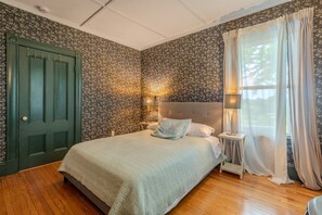 A bedroom with a bed, a nightstand, a lamp, a window with curtains, and a door.