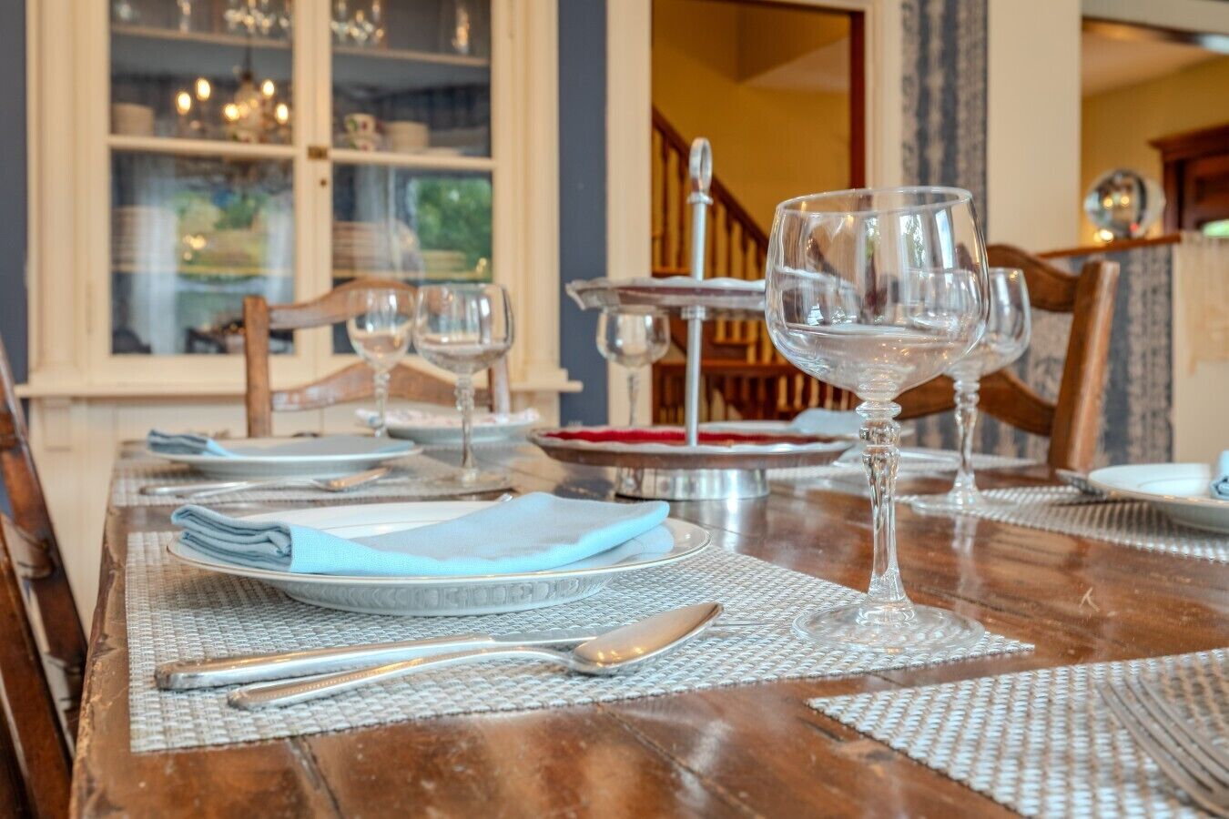Een gedekte eettafel met borden, bestek en wijnglazen, met zicht op een trap en een raam op de achtergrond.