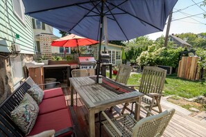 Un patio al aire libre con una mesa de madera, sillas, una parrilla y un paraguas rojo.