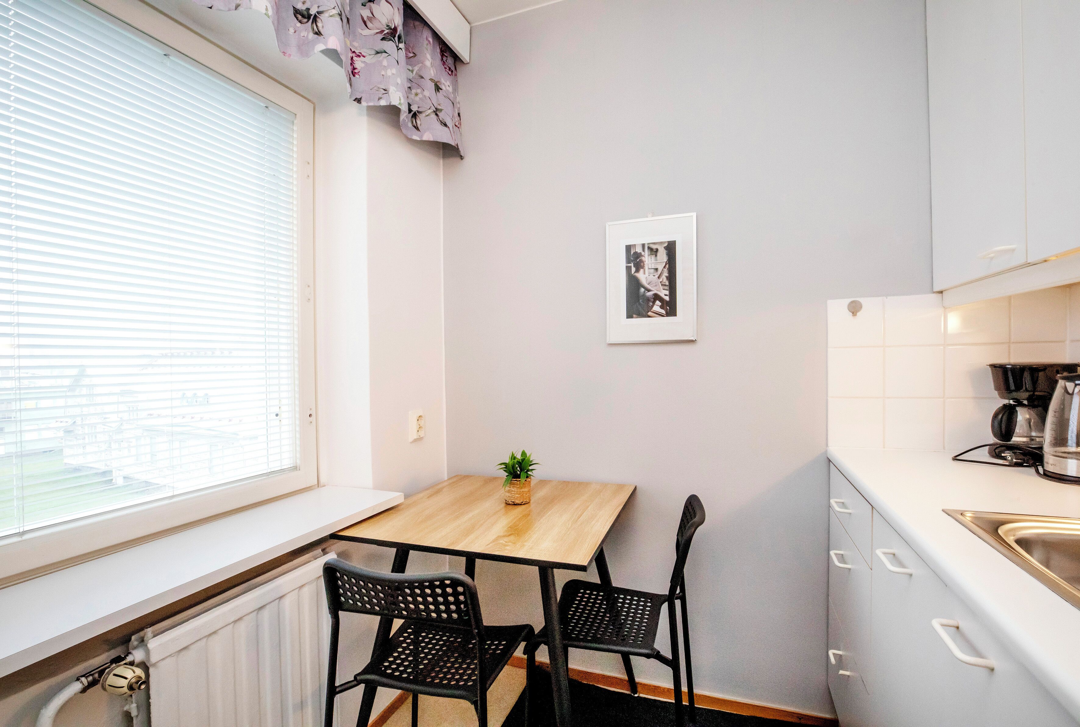 Et lille køkken med et vindue, et bord, to stole, et billede i ramme på væggen og en potteplante.