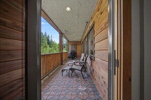 A covered outdoor balcony with a grill, chairs, and a view of the forest.