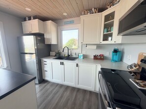 A modern kitchen with white cabinetry, a stainless steel refrigerator, a coffee bar, and a window with a view. - Beachfront Coastal Cottage with Harbour Views & Private Hot Tub-Mermaid  PEI (Mermaid)