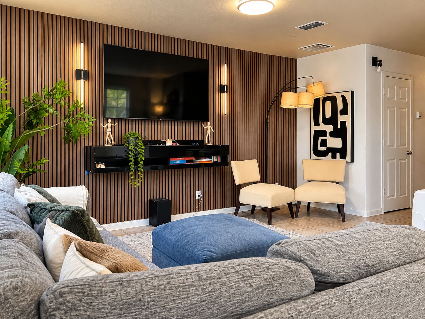 Een moderne woonkamer met een grote flatscreen-tv, een houten accentmuur, een zithoek met een bank en fauteuils, en een decoratieve plant.