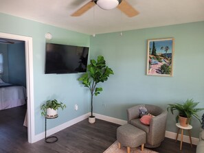 Ein Zimmer mit Deckenventilator, einem an der Wand befestigten Flachbildfernseher, einer großen Topfpflanze, einem grauen Sessel und einem Bild mit einer tropischen Szenerie.
