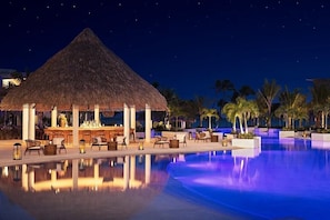 A nighttime resort pool area with a thatched-roof bar and lounge chairs. - Escape into luxury at Secrets® Cap Cana Resort & Spa, a AAA Five Diamond Resort! (Punta Cana)