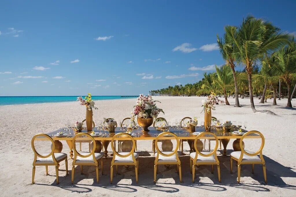 A beachside dining table set with chairs, a clear view of the ocean, and palm trees in the background.