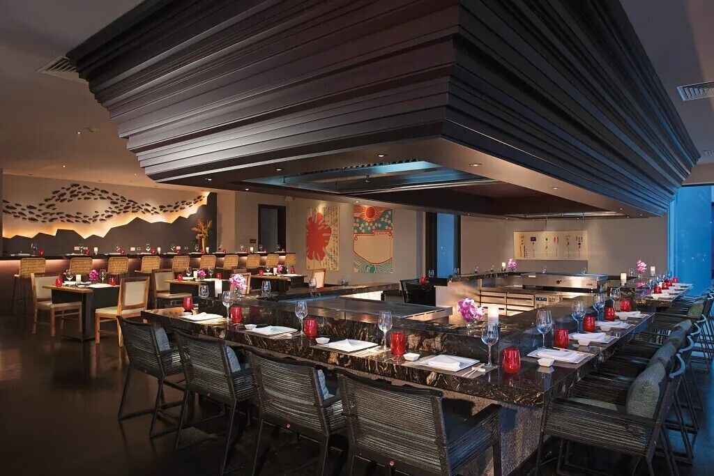 A modern restaurant with a long dining table, multiple chairs, and a high ceiling with a unique design.