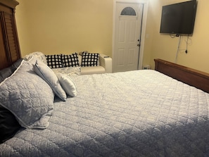 A bedroom with a large bed, a bedside table, a chair, a television mounted on the wall, and a door. - Your Ideal Stay Near the Stockyards (Fort Worth)