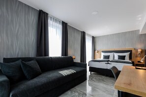 A modern hotel room with a dark-colored sofa, a bed with a wooden headboard, a wooden desk, and a marble floor. - Hotel Tříška (Stredocesky kraj)