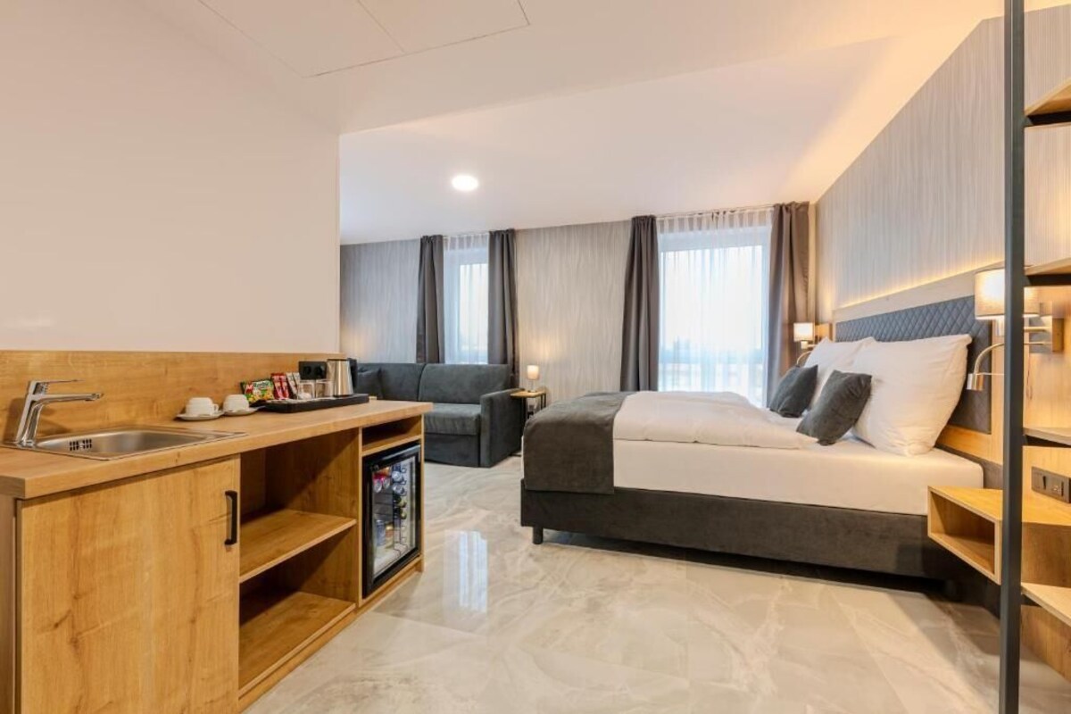 A modern hotel room with a bed, a sofa, a small kitchenette, and a bathroom.