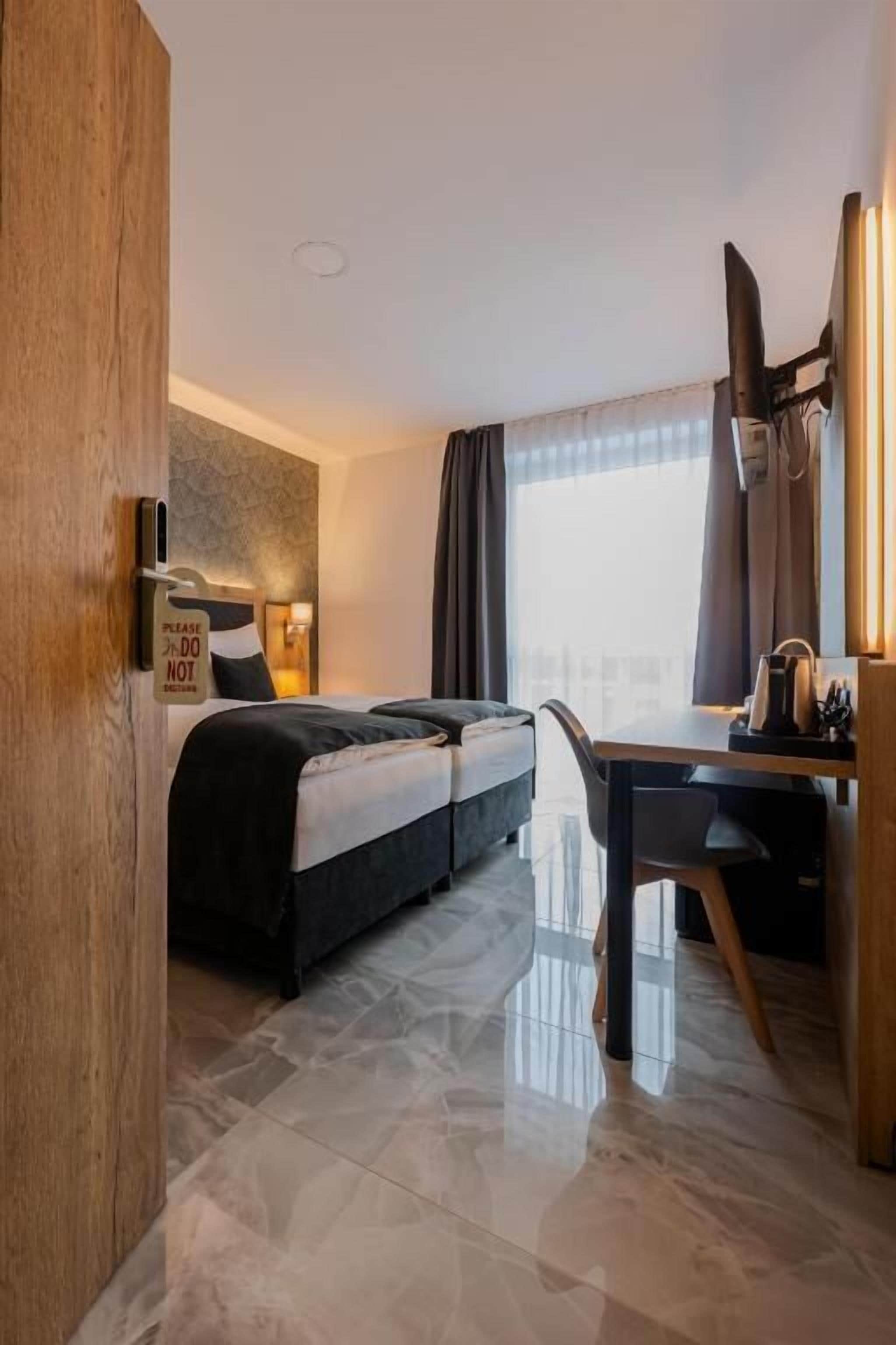Habitación de hotel con cama, escritorio, silla, ventana con cortinas y puerta.