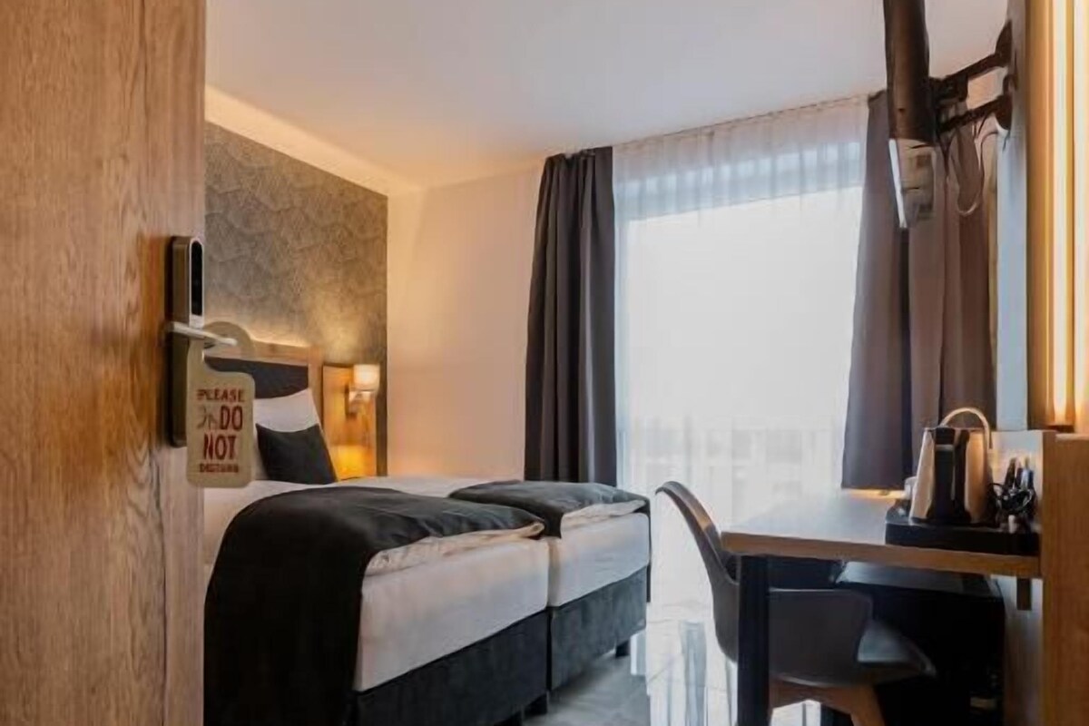 A hotel room with a bed, a desk, a chair, a window with curtains, and a door.