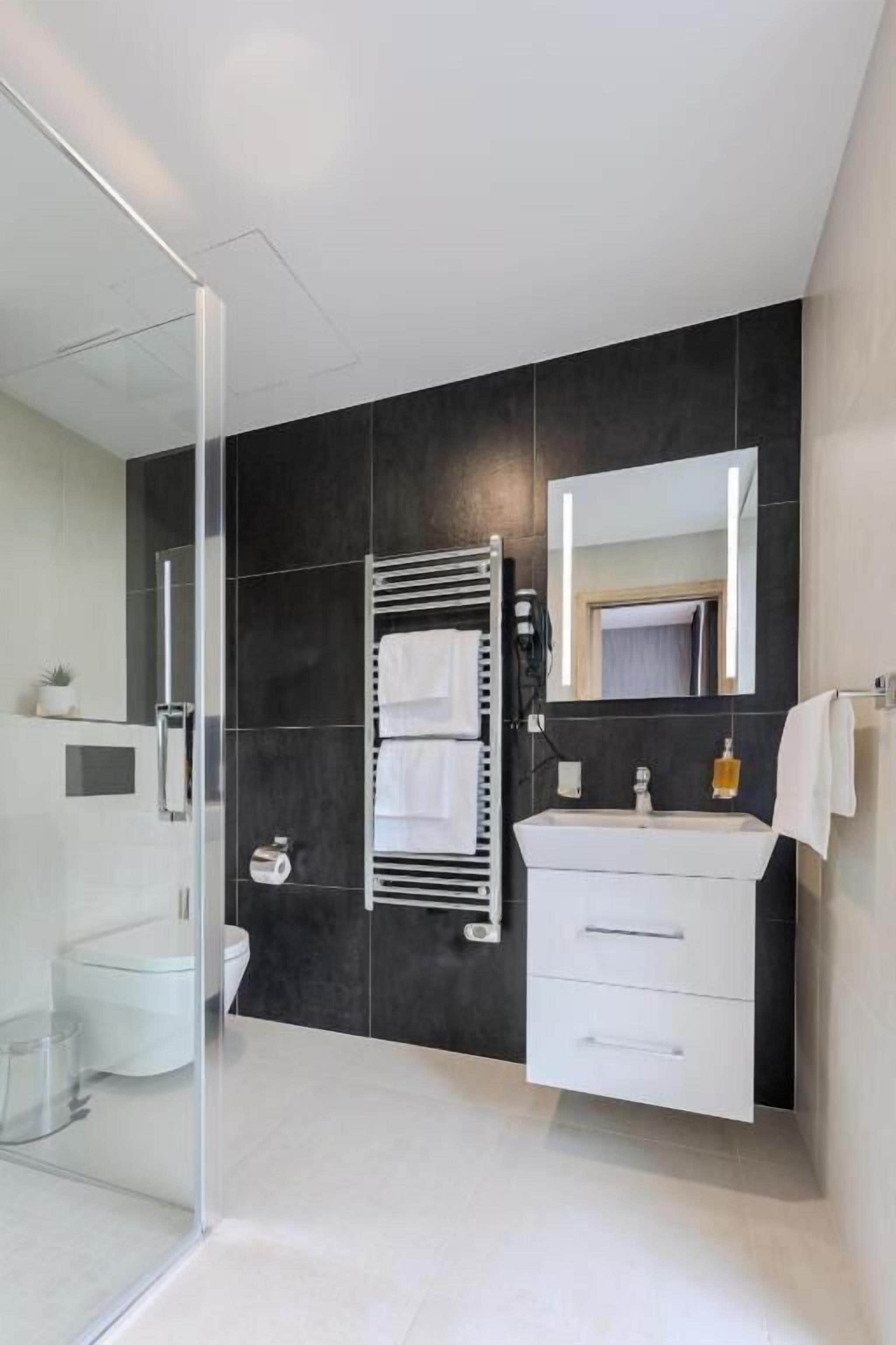 Un baño moderno con cabina de ducha de vidrio, un tocador blanco con espejo y una pared de azulejos oscuros.