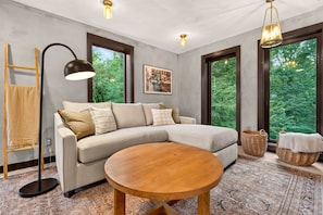 A modern living room with a sectional sofa, a round wooden coffee table, and large windows with a view of greenery. - The Prominence | Double Rooftop Lounge + Outdoor Movie (Tracy City)