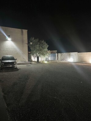 Un estacionamiento nocturno con un coche, un árbol y un edificio con entrada protegida.