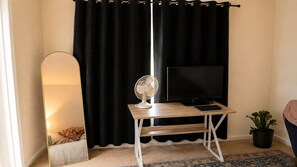 A room with a black curtain, a standing fan, a flat-screen TV, a small table, and a potted plant. - Guest Bedroom Furnished w/ Queen Bed, Firestick, TV, etc. (Springfield)