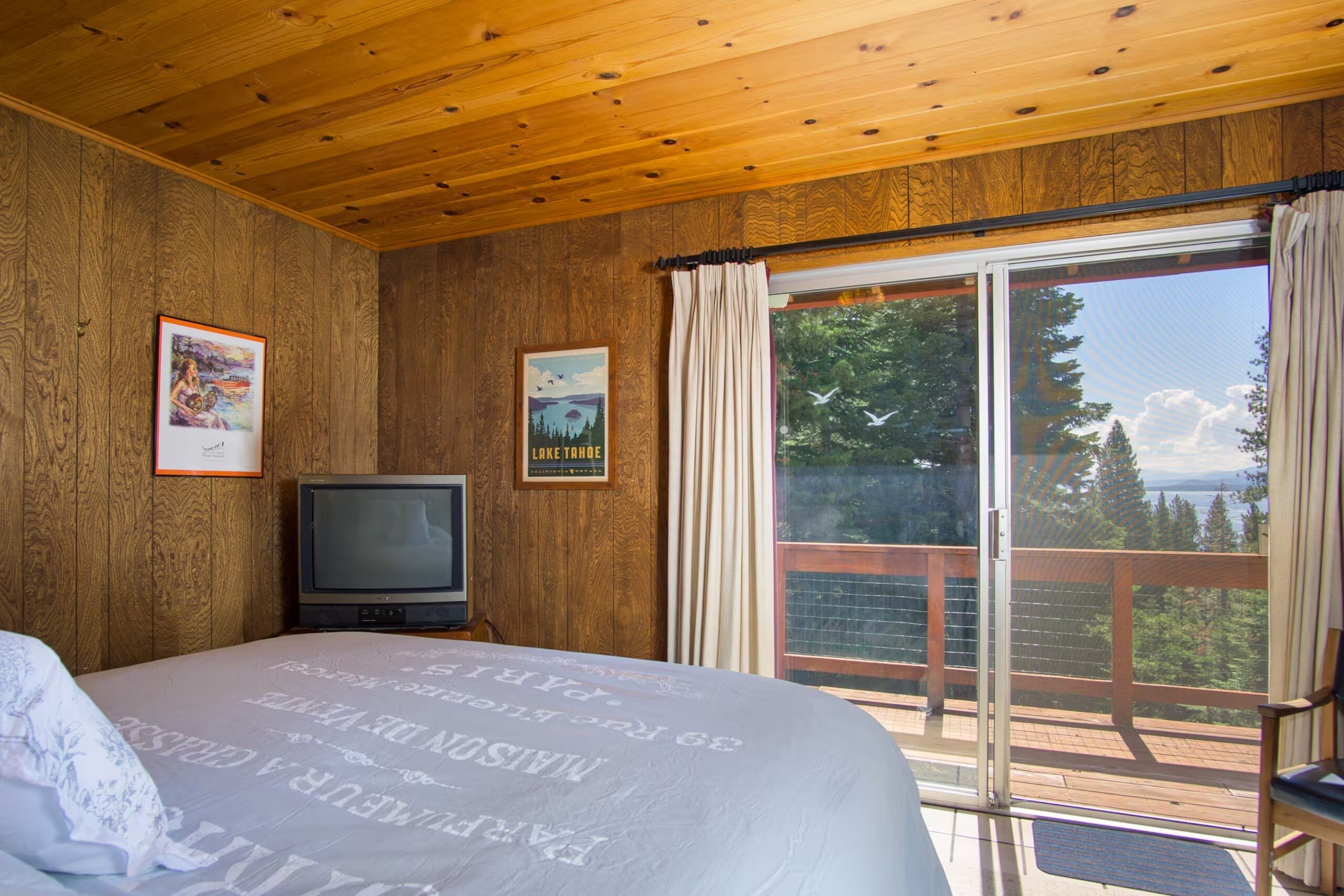 Une chambre dans un chalet en bois, équipée d’un lit, d’une télévision et d’un balcon offrant une vue sur les arbres et un lac.