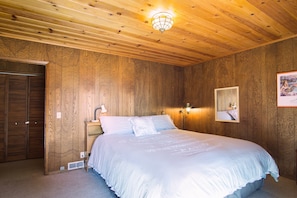 Un dormitorio con techo de madera, una cama con ropa de cama blanca, una mesita de noche, una lámpara de pared e imagen enmarcada.