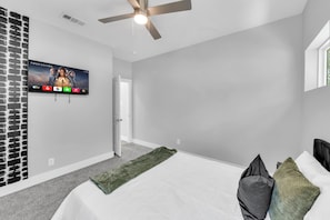A bedroom with a bed, a ceiling fan, a TV mounted on the wall, and a door leading to another room. - Modern 3BR Walk to Fair Park - EV Ready & Yard (Dallas)