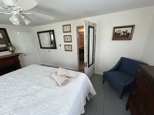 A bedroom with a bed, a chair, a dresser, a ceiling fan, a door, and a wall with framed pictures.