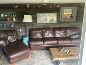 A living room with a brown leather sofa, a matching ottoman, a wooden coffee table, a lamp, and a wall-mounted shelf with decorative items. -  Farmhouse in Windsor. A scenic countryside escape. (Windsor)