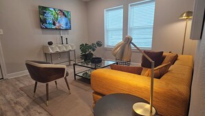 Uma sala de estar moderna com um sofá, uma mesa de centro, uma TV fixada na parede e uma janela com persianas.