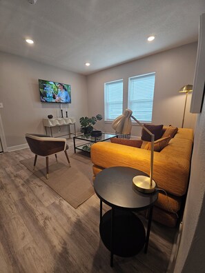 A modern living room with a sofa, a coffee table, a TV mounted on the wall, and a window with blinds. - Cozy 1-bedroom apartment in charming KC's 'Squire Park with in-unit washer/dryer (Kansas City)
