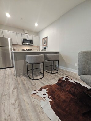 A modern kitchen with a stainless steel refrigerator, a microwave, and a sink. There are two bar stools and a cowhide rug on the floor. - Cozy 1-bedroom apartment in charming KC's 'Squire Park with in-unit washer/dryer (Kansas City)