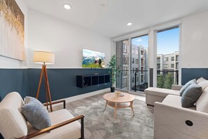 A modern living room with a sofa, armchair, and a small round table. There is a TV mounted on the wall and a potted plant on the floor. - Chic 1BR Stay | Sleeps 4 | Northern Liberties (Philadelphia)