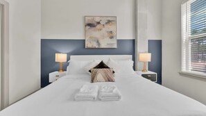 A neatly made bed with white linens and two pillows, flanked by bedside tables with lamps, in a room with a blue accent wall and a painting above the bed. - Modern Uptown 1BR | Sleeps 4 | Near Klyde Park (Dallas)