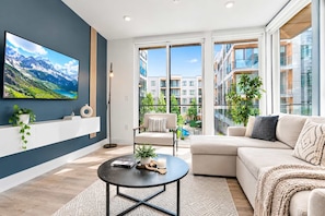A modern living room with a large flat-screen TV, a sofa, a coffee table, and a view of a building with balconies. - Fresh 1BD Near City Hall | Comfortably Sleeps 2 (Philadelphia)