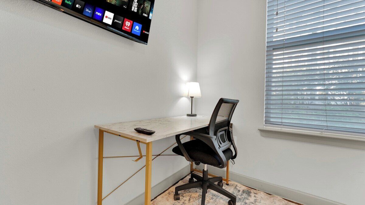 A room with a desk, chair, TV mounted on the wall, and a window with blinds.