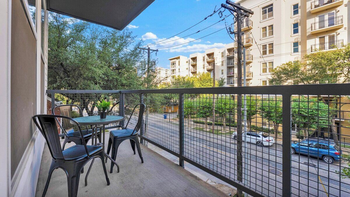 A balcony with a table and chairs, a view of a street with cars, and a building in the background.