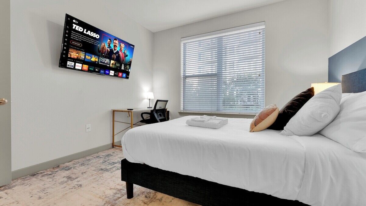 A hotel room with a bed, a desk, a TV mounted on the wall, and a window with blinds.