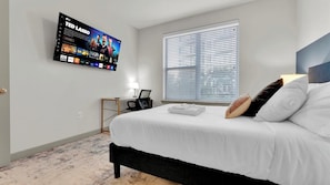 A hotel room with a bed, a desk, a TV mounted on the wall, and a window with blinds. - Uptown Escape 2BD | Sleeps 12 | Parking (Dallas)