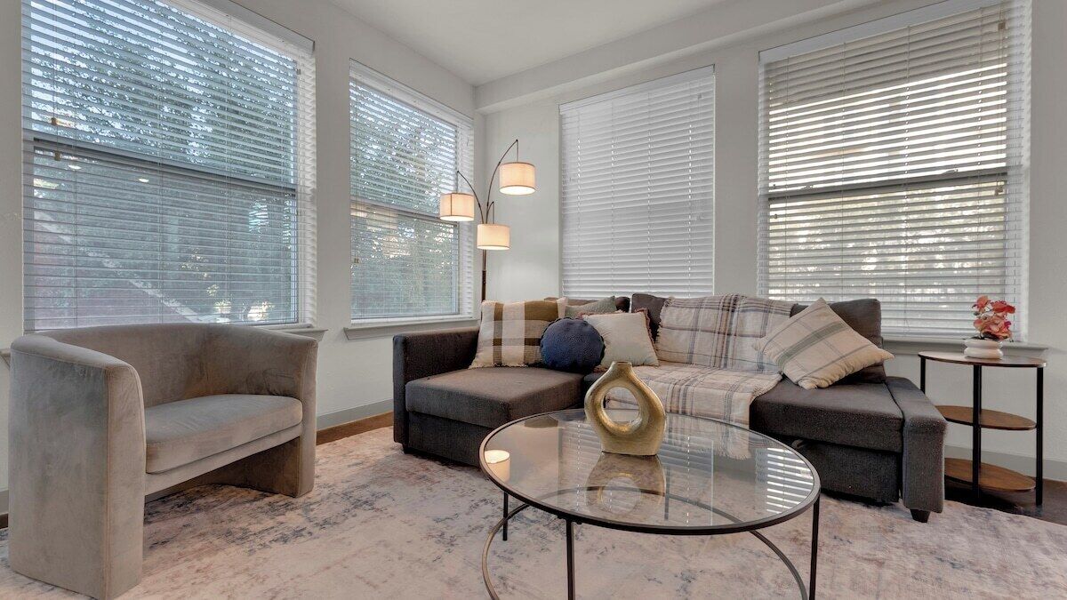 A living room with a sofa, armchair, and coffee table, large windows with blinds, and a vase on a side table.
