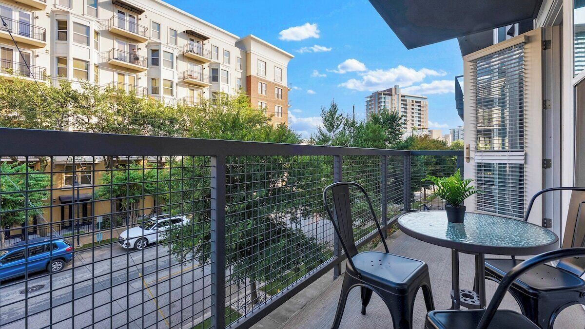 A balcony with a table and chairs, a potted plant, and a view of a street with cars and buildings.